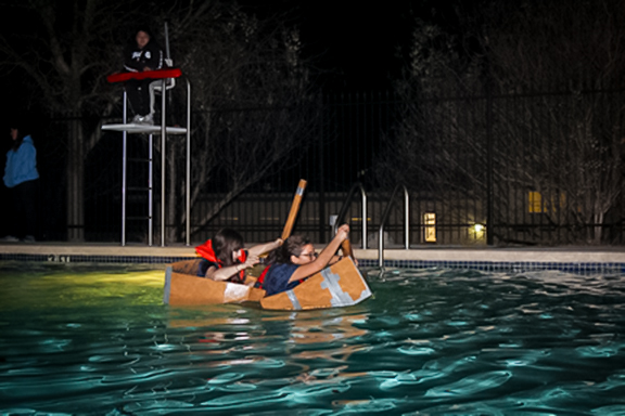 Girls paddling cardboard boat across pool at night