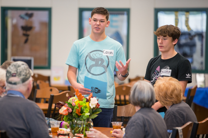 two male students standing and talking with senior adults at table