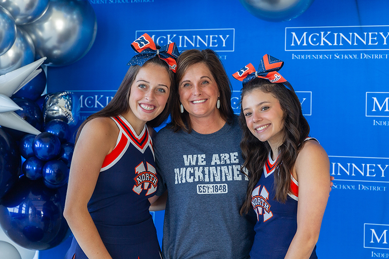 Wendy Dutton and two cheerleaders smiling for camera in front of MISD backdrop