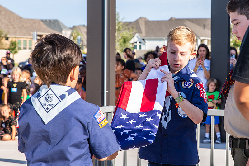 Cub scouts unfolding American flag by flagpole as crowd looks on 