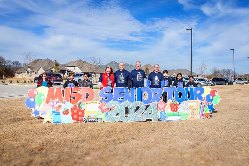 group of students and tour members standing behind large MISD senior tour sign in front of Frazier 