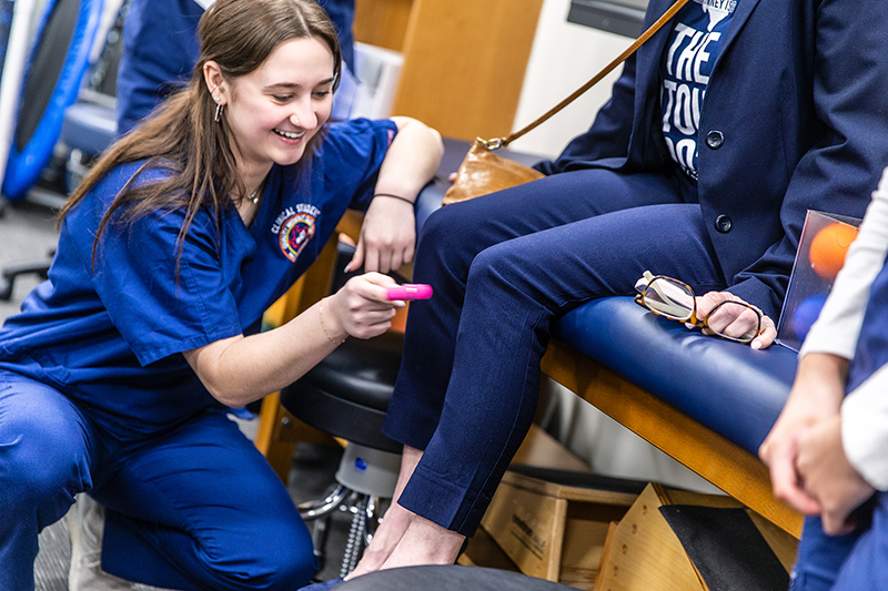 female sports medicine student checks a tour guest's reflexes using a medical knee tool as she laughs 