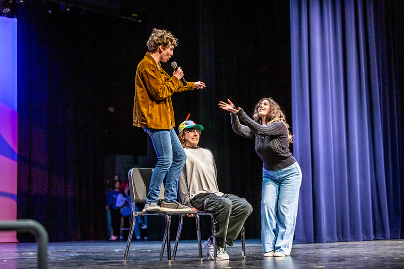 3 theater students perform a song from musical Big Fish as main character stands on a chair holding microphone and singing 